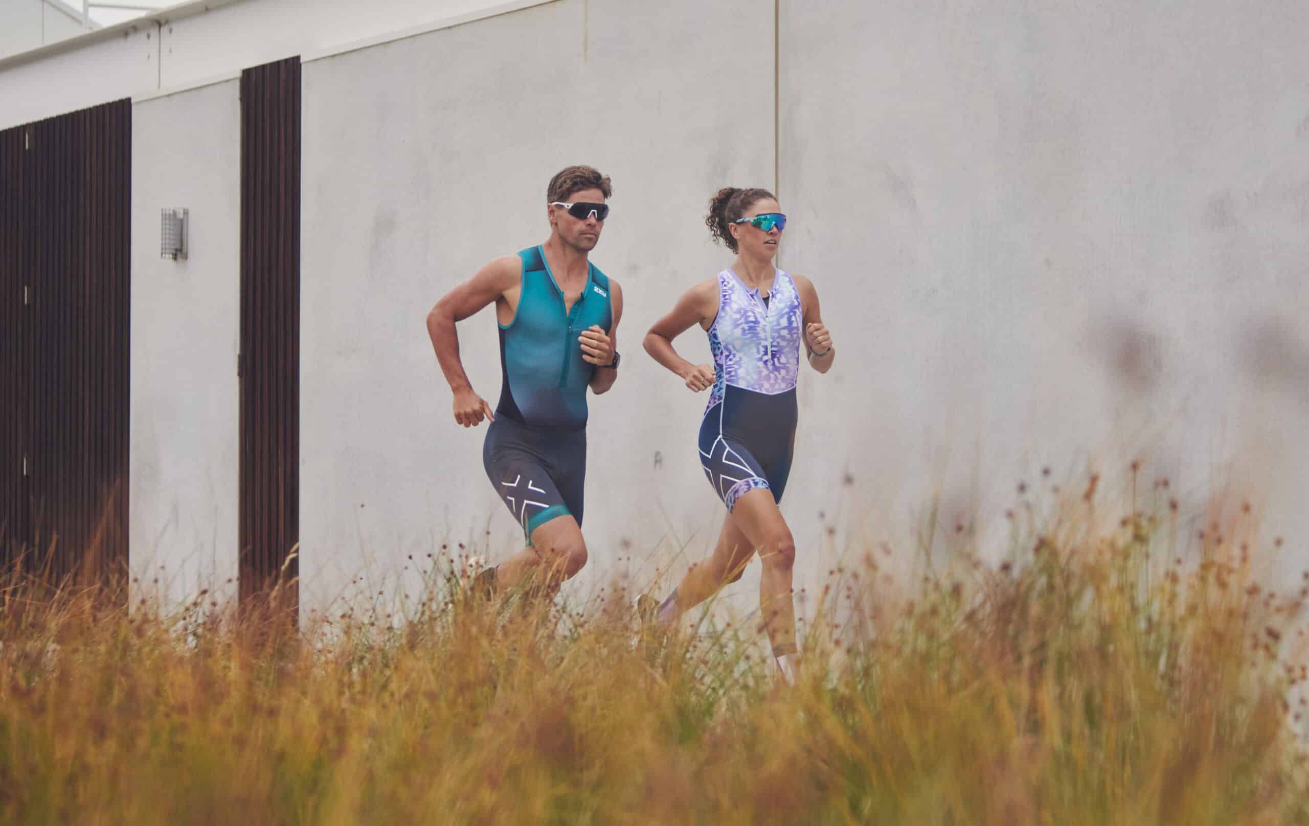 white man and women jogging next to white building long grass surrounds them man is wearing grey athletic shorts and a teal tank top woman is in grey spandex shorts and a white tank top