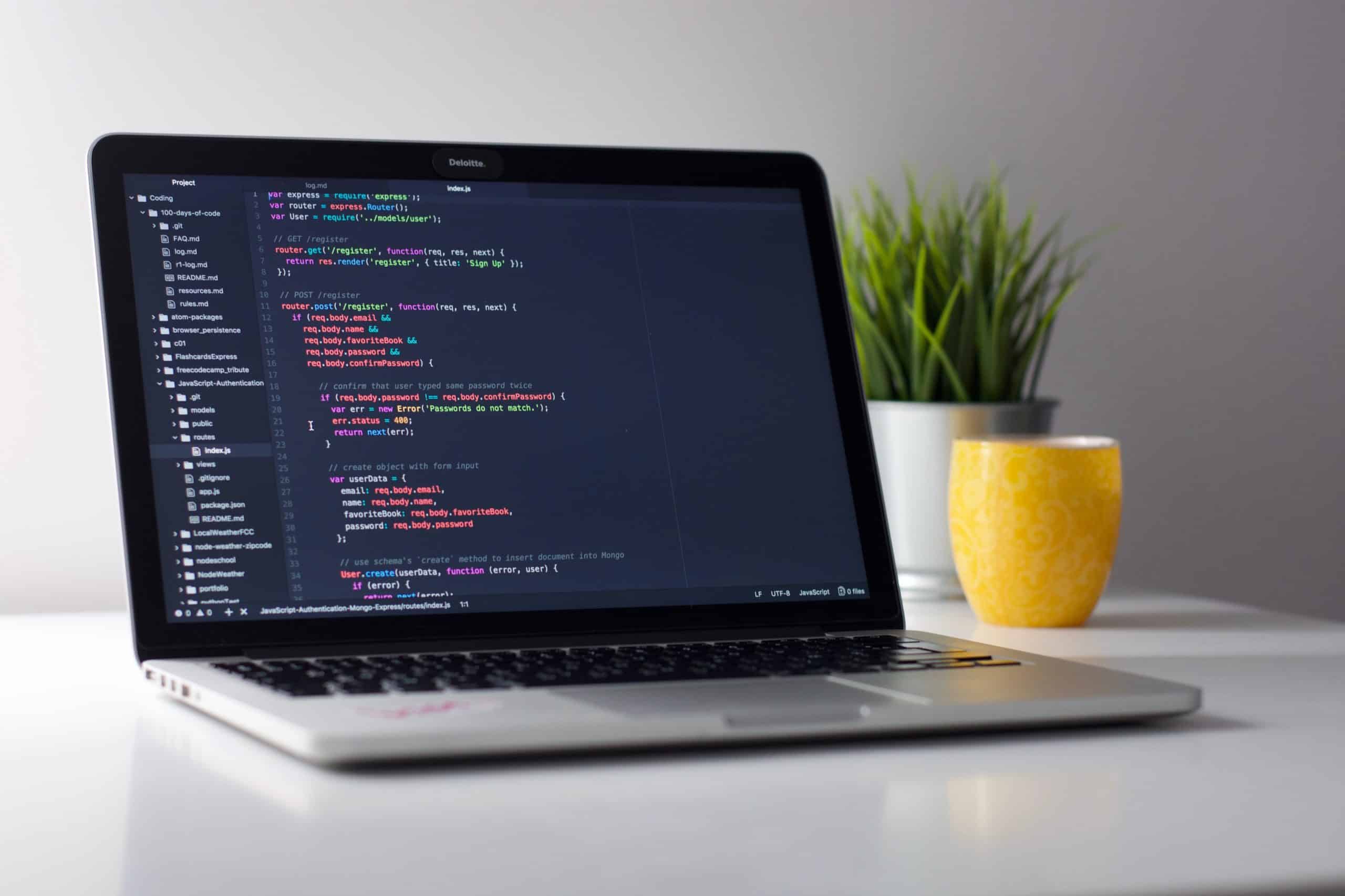 open laptop with black screen and colorful code; yellow coffee mug; white planter with green plant on table.