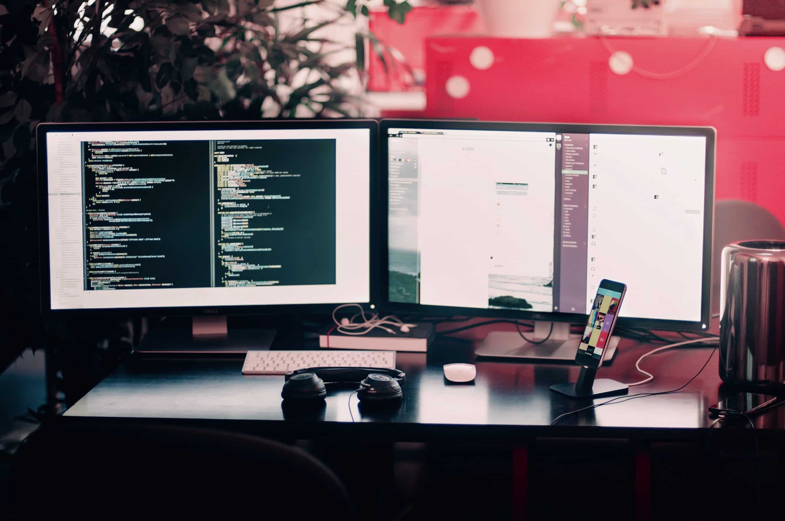 two computer screens with code; headphones and cell phone on desk; red wall in background with plant closer to desk