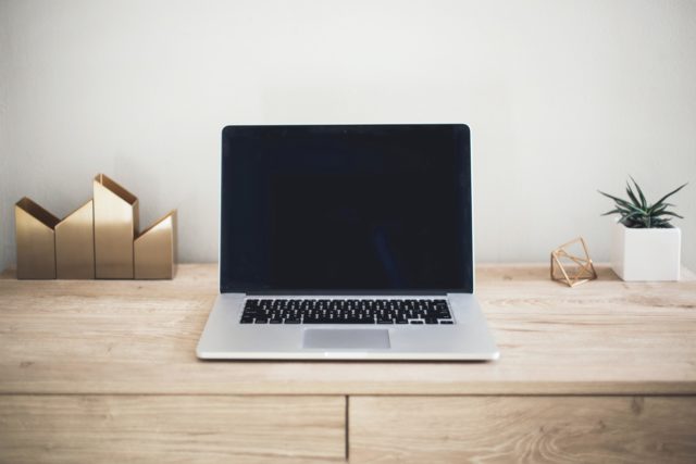 Laptop on light woodgrain desk with gold pencil holders, gold round modern figurine, and small plant in white planter. Valentine's Merchandising blogging visual. 