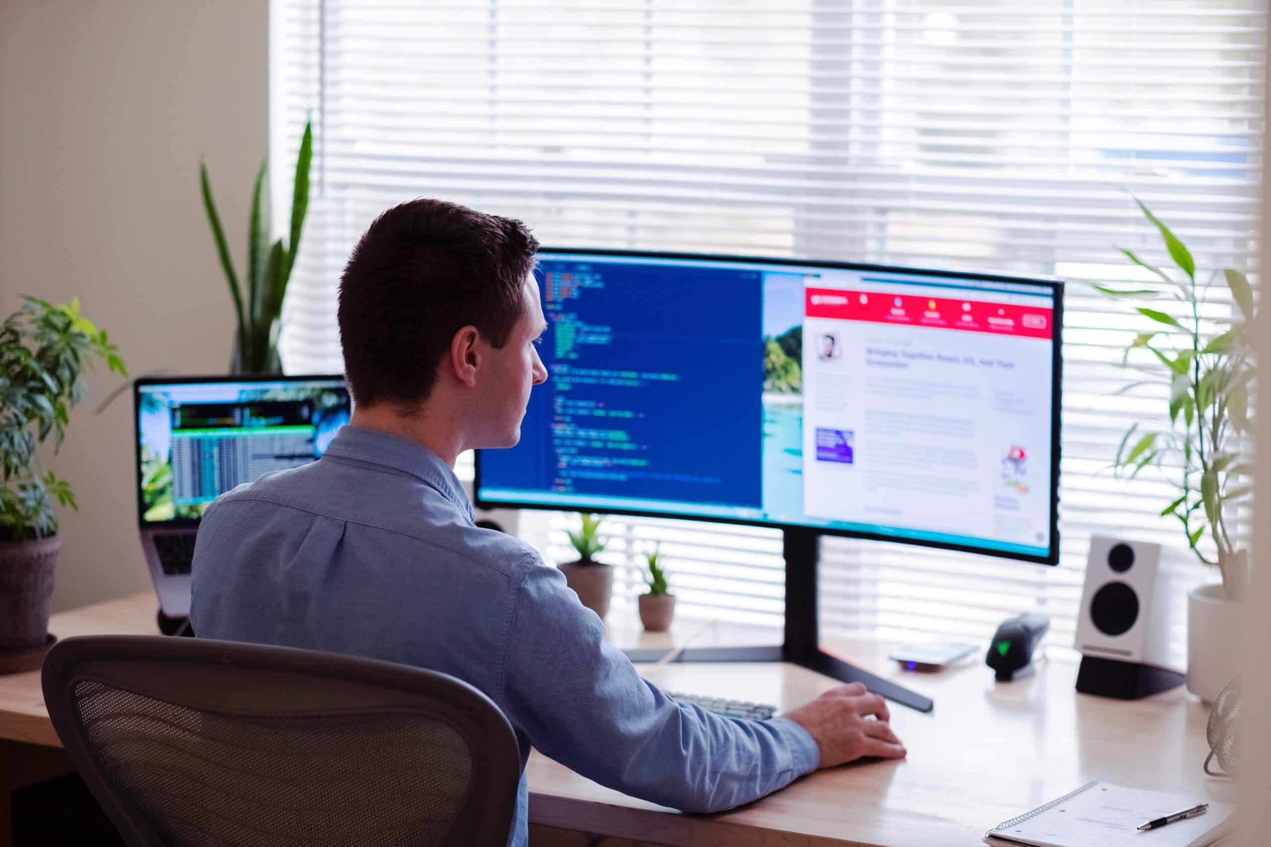 man at computer coding and reading an article; bright background with sunlight; three plants on desk; speaker on desk