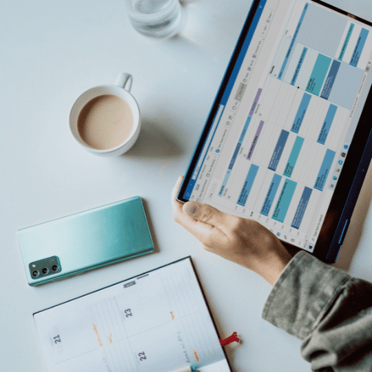 digital calendar on project management solution and a paper calendar book. Cell phone with blue-green case, coffee, hands holding tablet